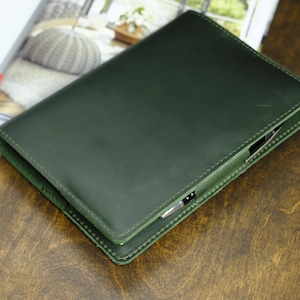 May include: A dark green leather journal with a pen holder, lying on a wooden surface. The journal is closed, with visible stitching around the edges. An open book with a red pen is in the background.