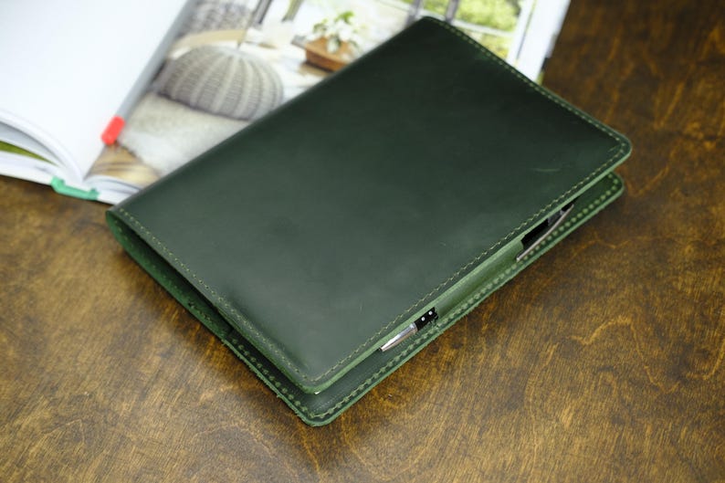 May include: A dark green leather journal with a pen holder, lying on a wooden surface. The journal is closed, with visible stitching around the edges. An open book with a red pen is in the background.