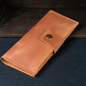 Może przedstawiać: Brązowy skórzany portfel z zapięciem na zatrzask. Portfel jest złożony na pół i ma klapkę, która zakrywa przód.