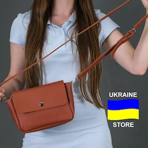 Może przedstawiać: Kobieta trzyma brązową skórzaną torebkę na ramię z srebrną klamrą i klapką. Torebka ma długi pasek i jest noszona na ramieniu. Kobieta ma na sobie jasnoniebieską koszulę polo i białą spódnicę. Obraz zawiera również tekst "UKRAINE STORE" i żółto-niebieską flagę.