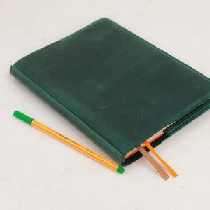Puede incluir: Un diario de cuero verde oscuro con un borde cosido, abierto para revelar marcadores de página coloridos. Un bolígrafo naranja y amarillo con punta verde descansa junto al diario. El diario es un artículo de papelería clásico.