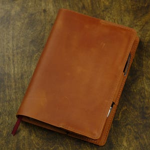 May include: A tan leather notebook cover with a smooth, matte finish and visible stitching along the edges. A thin, dark red fabric bookmark extends from the bottom left corner, and a silver pen is tucked into the side loop of the cover.