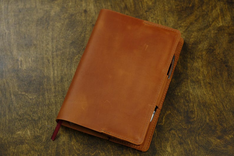 May include: A tan leather notebook cover with a smooth, matte finish and visible stitching along the edges. A thin, dark red fabric bookmark extends from the bottom left corner, and a silver pen is tucked into the side loop of the cover.