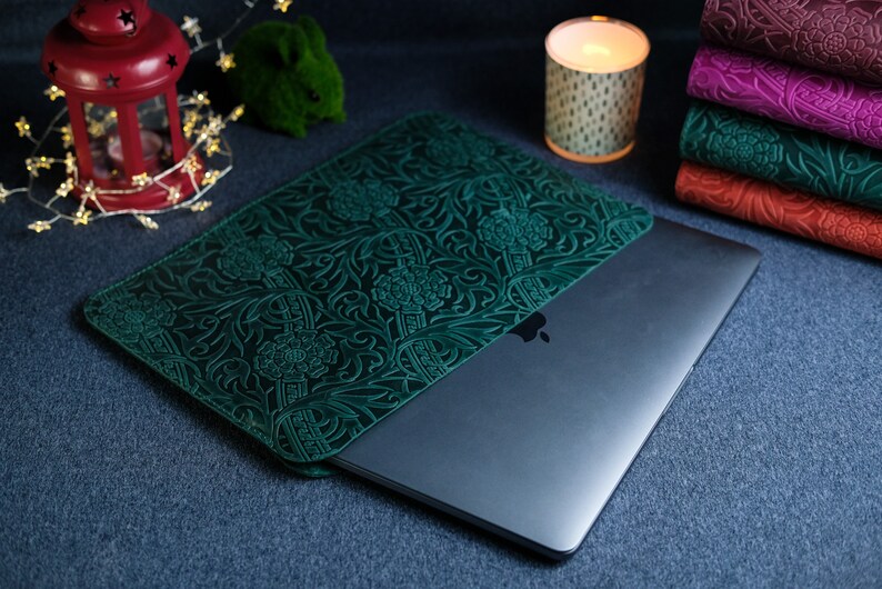 K&ouml;nnte beinhalten: Eine gr&uuml;ne Lederh&uuml;lle f&uuml;r Laptops mit einem komplizierten Blumenmuster. Die H&uuml;lle ist geschlossen und ein silberner Laptop ist teilweise im Inneren sichtbar.