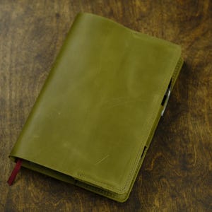 May include: A rectangular olive green leather notebook cover with visible stitching along the edges. The cover features a smooth, matte texture and a small, dark red fabric ribbon bookmark extending from the bottom left corner of the spine.