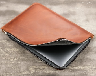 May include: A tan leather laptop sleeve with a silver zipper closure. The sleeve is partially open, revealing a grey felt interior lining and a silver metallic laptop partially inserted into the protective case.