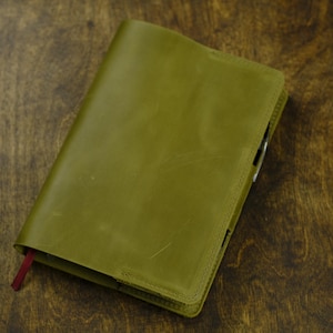 May include: Olive green leather journal with a red ribbon bookmark. The journal is closed and has a stitched border. The cover is slightly textured and appears to be made of high-quality leather. The journal is on a dark wood surface.