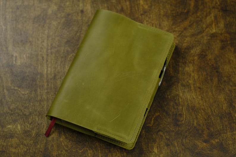May include: Olive green leather journal with a red ribbon bookmark. The journal is closed and has a stitched border. The cover is slightly textured and appears to be made of high-quality leather. The journal is on a dark wood surface.