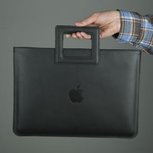 May include: Black leather laptop sleeve with a handle. The sleeve features a debossed Apple logo and a rectangular handle at the top. The sleeve is held by a person.
