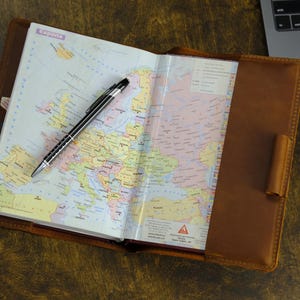 May include: An open brown leather notebook cover holding a planner with a printed map of Europe on the visible page. A black and silver ballpoint pen rests diagonally across the map. The leather cover features a pen loop on the right side.