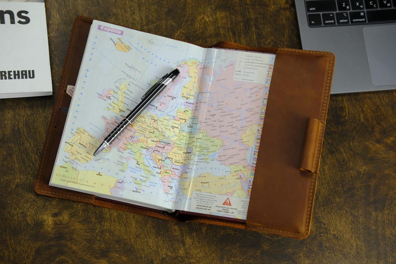 May include: An open brown leather notebook cover holding a planner with a printed map of Europe on the visible page. A black and silver ballpoint pen rests diagonally across the map. The leather cover features a pen loop on the right side.