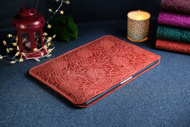 K&ouml;nnte beinhalten: Eine rote Lederh&uuml;lle f&uuml;r Laptops mit einem gepr&auml;gten Blumenmuster. Die H&uuml;lle liegt auf einer dunkelblauen Oberfl&auml;che.