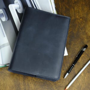 May include: A dark blue leather journal lies open on a wooden surface, next to a black pen with silver accents and a pencil with a red eraser. The journal is closed, with a stitched border. A notebook and a magazine are in the background.