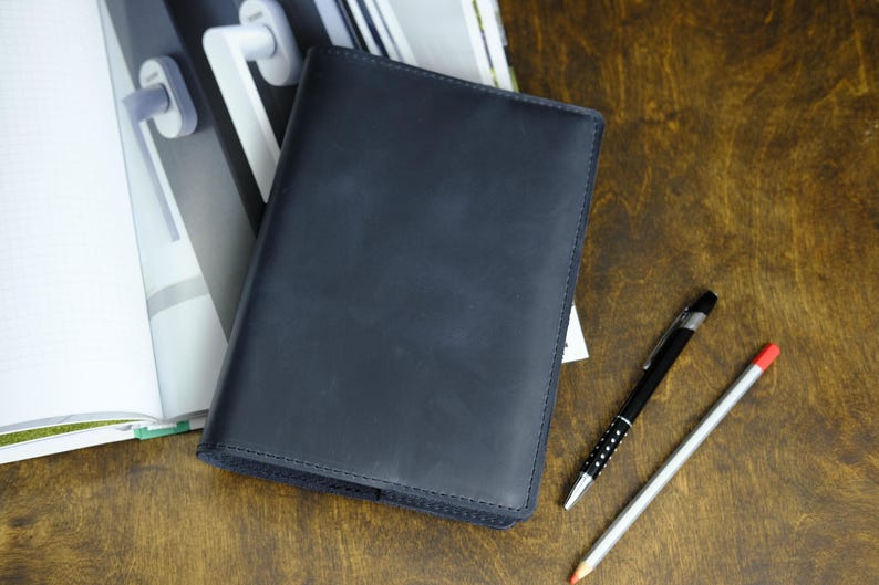 May include: A dark blue leather journal lies open on a wooden surface, next to a black pen with silver accents and a pencil with a red eraser. The journal is closed, with a stitched border. A notebook and a magazine are in the background.