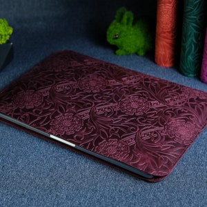 K&ouml;nnte beinhalten: Eine dunkelrote Laptoptasche aus Leder mit einem gepr&auml;gten Blumenmuster. Die Tasche liegt auf einer blauen Oberfl&auml;che.