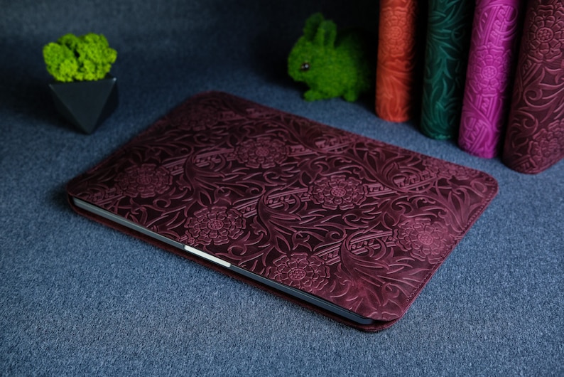 K&ouml;nnte beinhalten: Eine dunkelrote Laptoptasche aus Leder mit einem gepr&auml;gten Blumenmuster. Die Tasche liegt auf einer blauen Oberfl&auml;che.