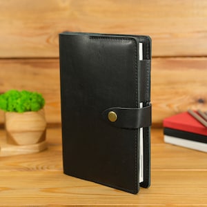 May include: A black leather journal with a strap closure and a brass button. The journal is closed and standing upright. Other items on the wooden surface include a small wooden planter with green moss, a pen, and a stack of notebooks with pencils.