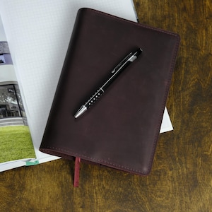 May include: A dark burgundy leather-bound journal with a red ribbon bookmark and a black pen with silver accents. The journal rests on a wooden surface next to a notebook and a laptop.
