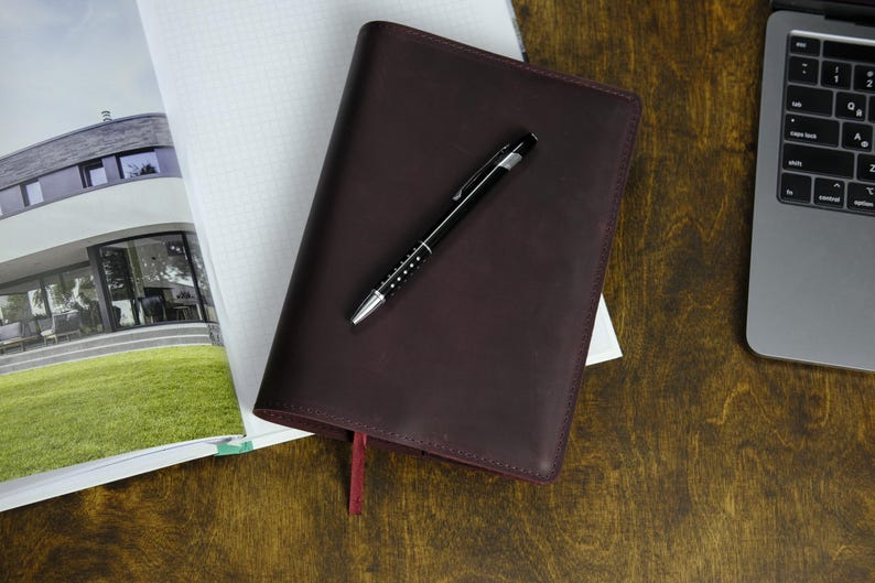 May include: A dark burgundy leather-bound journal with a red ribbon bookmark and a black pen with silver accents. The journal rests on a wooden surface next to a notebook and a laptop.