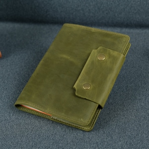 Może przedstawiać: Zielony skórzany notatnik w kolorze oliwkowym z pasującą klapką zamykającą, zabezpieczoną dwoma zatrzaskami w kolorze mosiądzu. Notatnik jest prostokątny i wygląda na ręcznie robiony. W tle widoczny jest stos notatników w różnych kolorach.