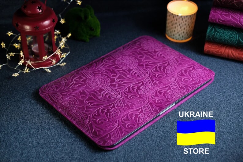 K&ouml;nnte beinhalten: Eine violette Lederh&uuml;lle f&uuml;r Laptops mit einem gepr&auml;gten Blumenmuster. Die H&uuml;lle ist geschlossen und die Kanten sind sichtbar. Der Hintergrund ist eine dunkelblaue Oberfl&auml;che. Der Text "UKRAINE STORE" und die ukrainische Flagge sind in der unteren rechten Ecke des Bildes sichtbar.