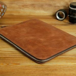 May include: A rectangular, tan-colored leather document sleeve or laptop case featuring a smooth, matte finish and a silver-toned metal zipper closure that runs along two sides of the item, providing a secure opening for the main compartment.