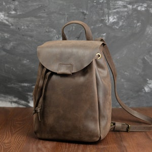 Op de afbeelding: Een bruine leren rugzak met een trekkoord sluiting en een klep. De rugzak heeft een enkele band en een gespsluiting.