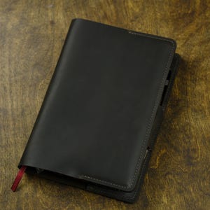 May include: A dark charcoal gray leather notebook cover with visible stitching along the edges. The cover features a smooth matte finish and a single thin red fabric bookmark ribbon extending from the bottom left corner of the rectangular object.