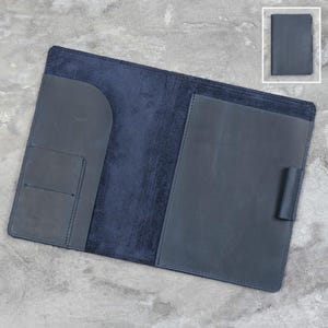 Può includere: Una copertina per taccuino in pelle blu scuro con interno aperto, dotata di una tasca verticale sul lato sinistro e di una tasca a tutta lunghezza sul lato destro, comprensiva di un passante per penna. Un piccolo riquadro mostra la copertina chiusa.