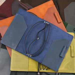 Puede incluir: Una funda de cuero azul para cuaderno con acabado mate, dos bolsillos interiores con bordes angulados, un portalápices en el lado derecho y un sistema de cordón elástico negro que pasa por ojales metálicos para asegurar los insertos.