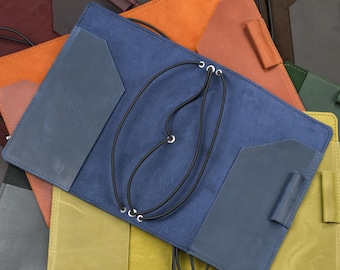 May include: A blue leather notebook cover featuring a matte finish, two interior pockets with angled edges, a pen loop on the right side, and a black elastic cord system threaded through metal eyelets to secure inserts within the cover.