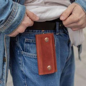 May include: A rectangular brown leather key holder featuring two silver-toned metal snap buttons for closure. The key holder is attached to a black belt loop on blue denim jeans, showcasing a smooth texture and visible stitching along the edges.