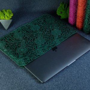 K&ouml;nnte beinhalten: Eine gr&uuml;ne Lederh&uuml;lle f&uuml;r Laptops mit einem Blumenmuster. Die H&uuml;lle ist geschlossen und ein Laptop ist teilweise im Inneren sichtbar.