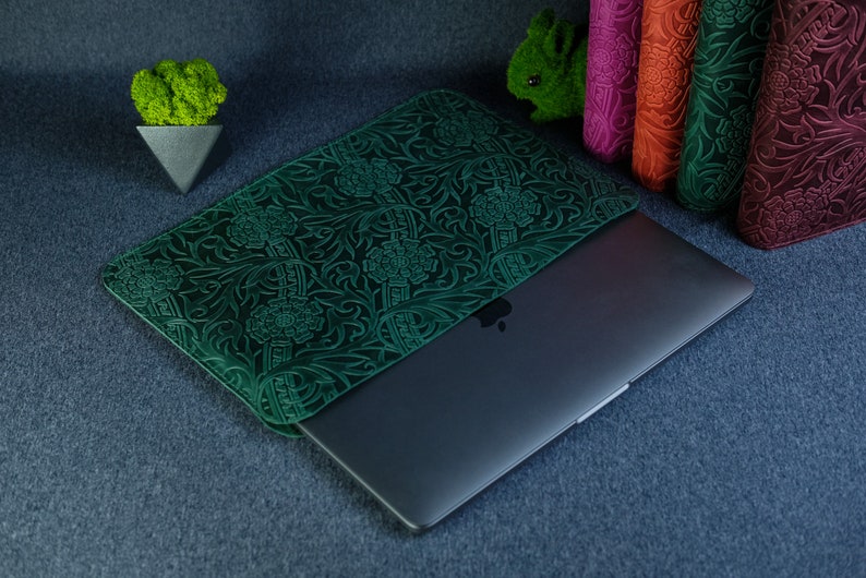 K&ouml;nnte beinhalten: Eine gr&uuml;ne Lederh&uuml;lle f&uuml;r Laptops mit einem Blumenmuster. Die H&uuml;lle ist geschlossen und ein Laptop ist teilweise im Inneren sichtbar.