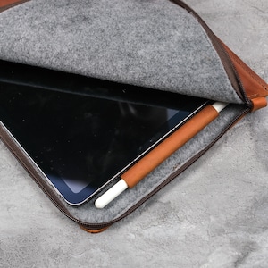 May include: A brown leather tablet sleeve featuring a gray felt interior lining, a dark brown zipper closure, and an integrated brown leather pen loop holding a white stylus. The sleeve is partially open, revealing a black tablet screen inside.
