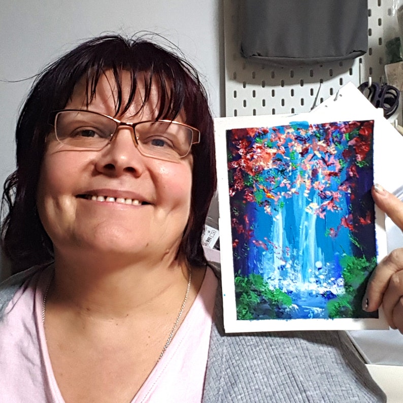 May include: A woman is holding a painting of a waterfall in a forest. The painting is on a white canvas and has a blue, green, and pink color scheme.
