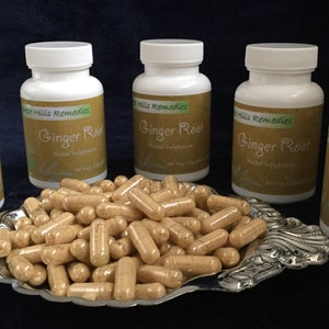 May include: Five white bottles of ginger root capsules with green labels and the text "Green Hills Remedies" and "Ginger Root" on them. The bottles are arranged in a row on a blue surface. A silver tray with a pile of ginger root capsules is in front of the bottles.