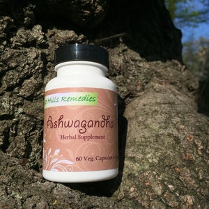 May include: A white bottle with a black lid labeled "Ashwagandha Herbal Supplement 60 Veg. Capsules" by "The Hills Remedies". The bottle is sitting on a tree trunk.