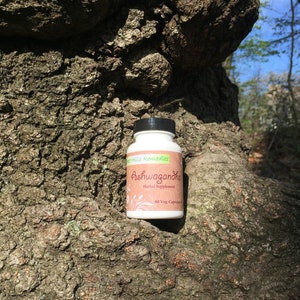 May include: A white bottle with a black lid and green label. The label says "Green Hills Remedies Ashwagandha Herbal Supplement 60 Veg. Capsules". The bottle is sitting on a tree trunk.