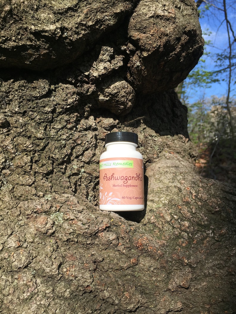 May include: A white bottle with a black lid and green label. The label says "Green Hills Remedies Ashwagandha Herbal Supplement 60 Veg. Capsules". The bottle is sitting on a tree trunk.