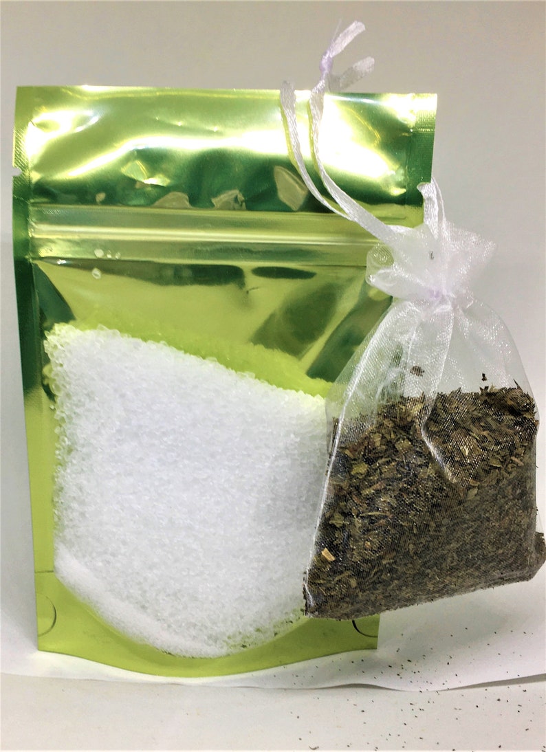 May include: A green resealable pouch with a white zipper contains white bath salts. A white mesh bag with a lavender ribbon contains dried herbs.