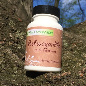 May include: A white bottle with a black lid labeled "Forest Hills Remedies Ashwagandha Herbal Supplement 60 Veg. Capsules". The bottle is sitting on a tree trunk.