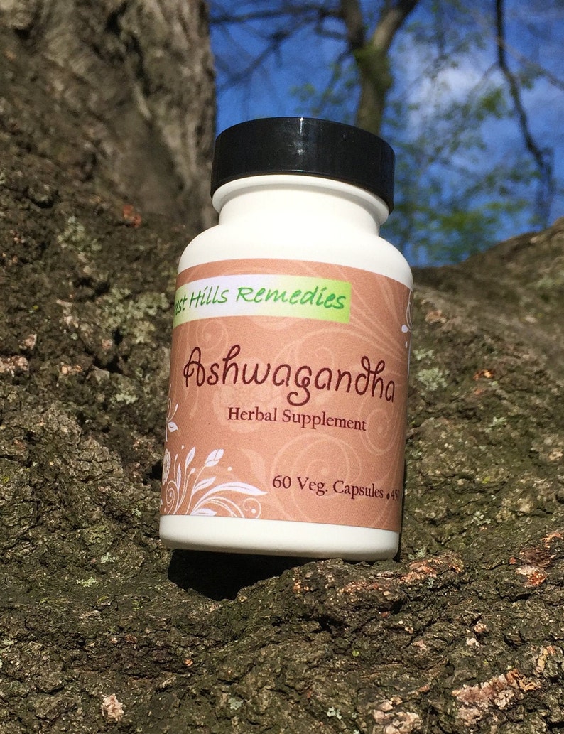 May include: A white bottle with a black lid labeled "Forest Hills Remedies Ashwagandha Herbal Supplement 60 Veg. Capsules". The bottle is sitting on a tree trunk.
