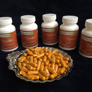 Op de afbeelding: Vijf witte flessen met kurkumacapsules met het label "Sweet Hills Remedies Kurkuma Kruiden Supplement 60 Vegetarische Capsules" en een kom met kurkumacapsules.