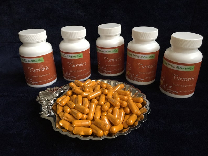 Op de afbeelding: Vijf witte flessen met kurkumacapsules met het label "Sweet Hills Remedies Kurkuma Kruiden Supplement 60 Vegetarische Capsules" en een kom met kurkumacapsules.