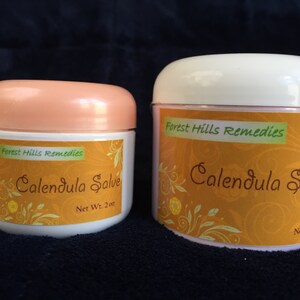 May include: Two white jars with orange labels. The labels read "Forest Hills Remedies Calendula Salve". The smaller jar is labelled "Net Wt. 2 oz" and the larger jar is labelled "Net Wt. 4 oz".