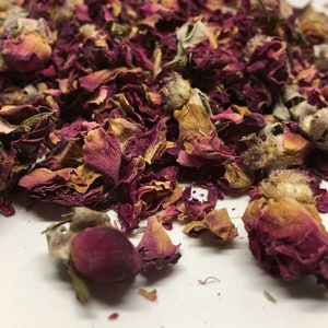 May include: Dried rose petals and buds in shades of pink, red, and brown. The petals are loose and scattered on a white surface.