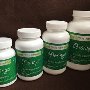 May include: Four white bottles of Moringa herbal supplement, labeled with green and white text. The bottles are arranged in a row from largest to smallest, with the largest bottle on the right. The bottles contain 400, 200, 160, and 90 vegetarian capsules.