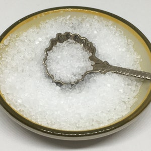 May include: A small white ceramic dish filled with white salt crystals. A silver spoon with a decorative shell-shaped bowl is filled with salt and rests on top of the salt in the dish.