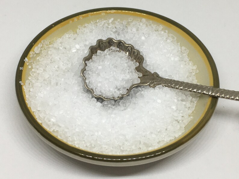 May include: A small white ceramic dish filled with white salt crystals. A silver spoon with a decorative shell-shaped bowl is filled with salt and rests on top of the salt in the dish.